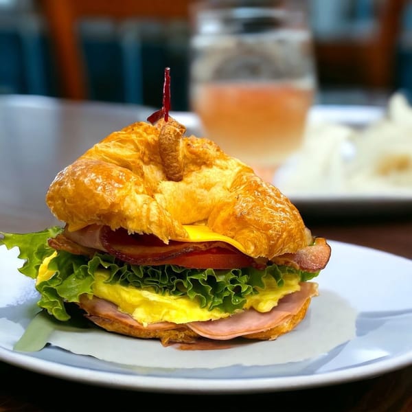 Croissant sandwich with ham, egg, cheese, bacon, lettuce, and tomato on a white plate.