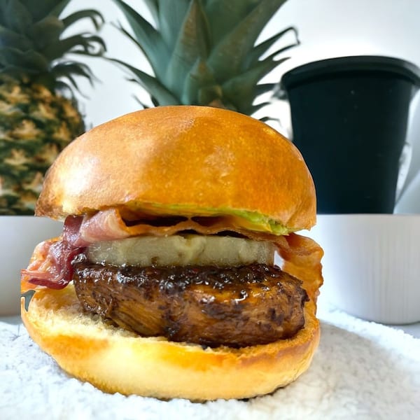 A gourmet burger with bacon, pineapple, and a dark glazed patty on a brioche bun.