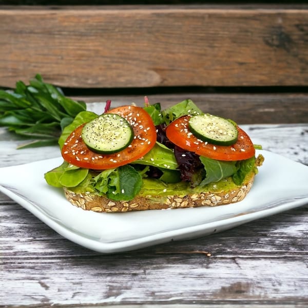 Avocado toast with lettuce, tomato slices, cucumber, and sesame seeds on a white plate.